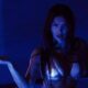 Megan Fox em luz azul, com biquíni prateado e gotículas caindo da mão estendida, olhar intenso