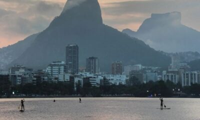 Pessoas em stand-up paddle em lago. Skyline urbano, montanhas ao fundo, céu nublado