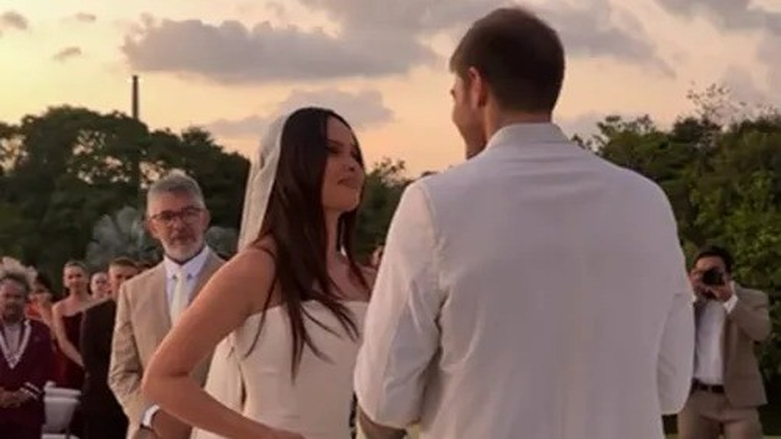 Juliette em vestido de noiva olhando para Kaique Cerveny em cerimônia com convidados ao fundo
