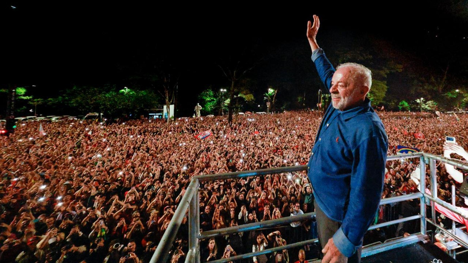 Presidente Lula no palco, acenando para uma vasta multidão com celulares acesos, à noite