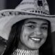 Mulher jovem sorrindo, usando chapéu de cowboy decorado e gargantilha, em preto e branco