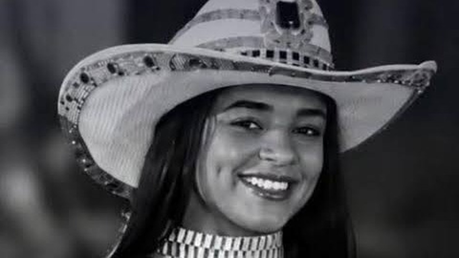Mulher jovem sorrindo, usando chapéu de cowboy decorado e gargantilha, em preto e branco
