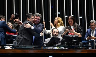 Pessoas em plenário, homens abraçam, outros usam celulares, um com câmera. "SENADO FEDERAL" em tela