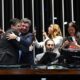 Pessoas em plenário, homens abraçam, outros usam celulares, um com câmera. "SENADO FEDERAL" em tela