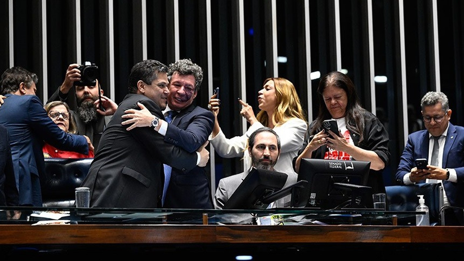 Pessoas em plenário, homens abraçam, outros usam celulares, um com câmera. "SENADO FEDERAL" em tela