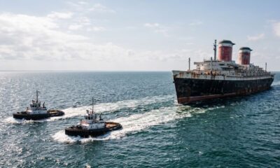 Dois rebocadores puxam um grande navio de passageiros enferrujado com duas chaminés em mar aberto