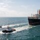 Dois rebocadores puxam um grande navio de passageiros enferrujado com duas chaminés em mar aberto