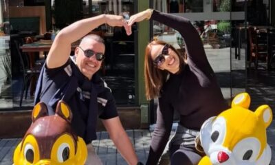 Um casal sorridente, de óculos escuros, formando um coração com as mãos, com estátuas de esquilos