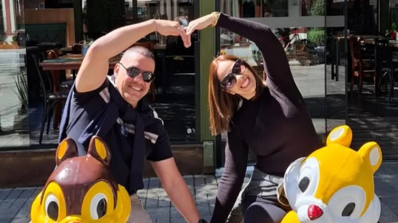 Um casal sorridente, de óculos escuros, formando um coração com as mãos, com estátuas de esquilos
