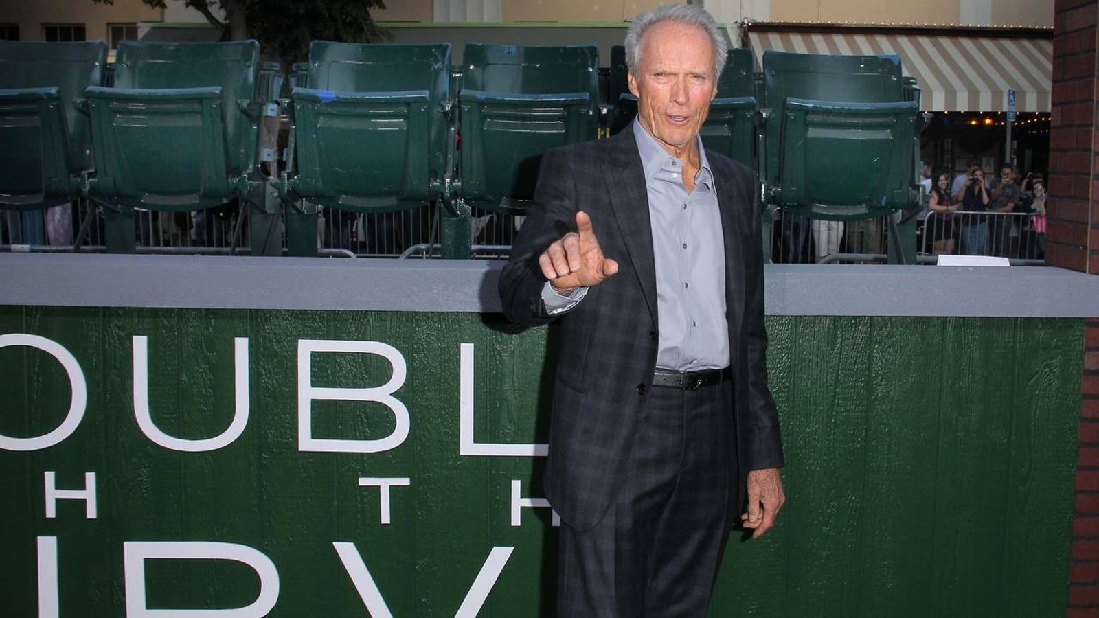 Clint Eastwood, terno xadrez, camisa cinza, apontando. Fundo: letras "DUBL HTH IDV", assentos