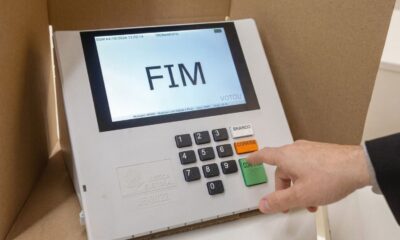 Mão pressiona 'CONFIRMA' em urna eletrônica branca exibindo 'FIM' e 'TREINAMENTO' na tela.