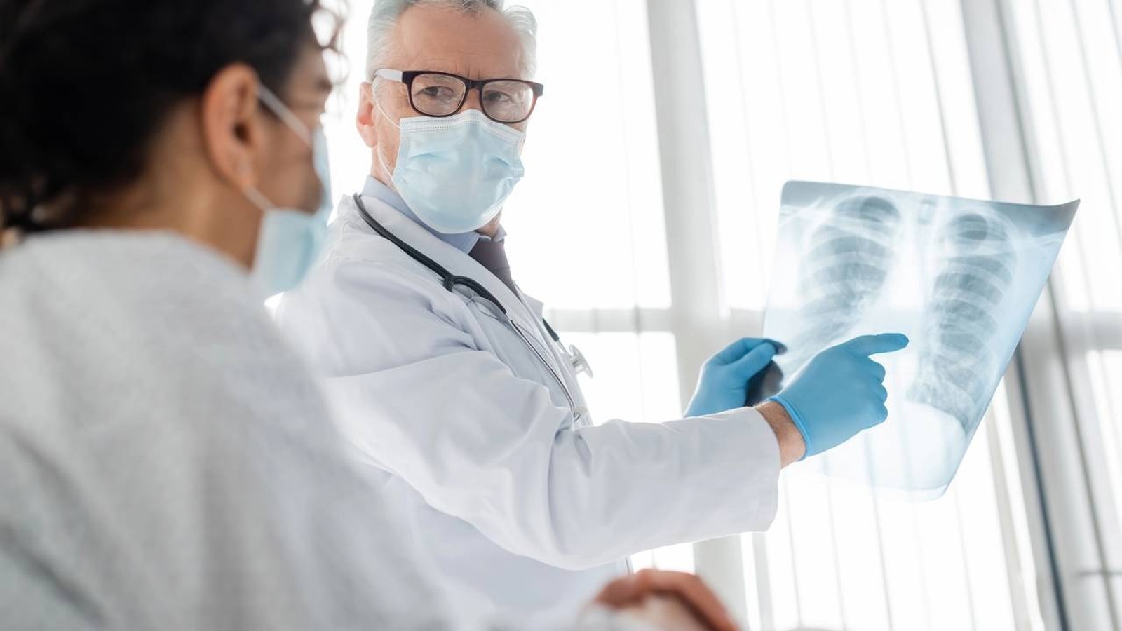 Médico de máscara e luvas aponta radiografia de tórax para paciente