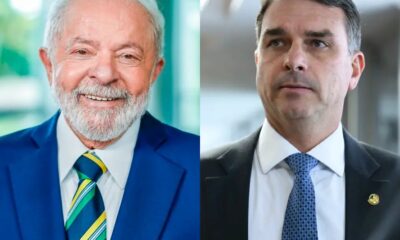 Lula sorridente de terno azul e Flávio Bolsonaro sério de terno escuro, lado a lado