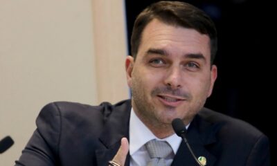 Flávio Bolsonaro de terno e gravata, gesticulando com a mão direita em frente a um microfone
