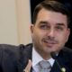 Flávio Bolsonaro de terno e gravata, gesticulando com a mão direita em frente a um microfone