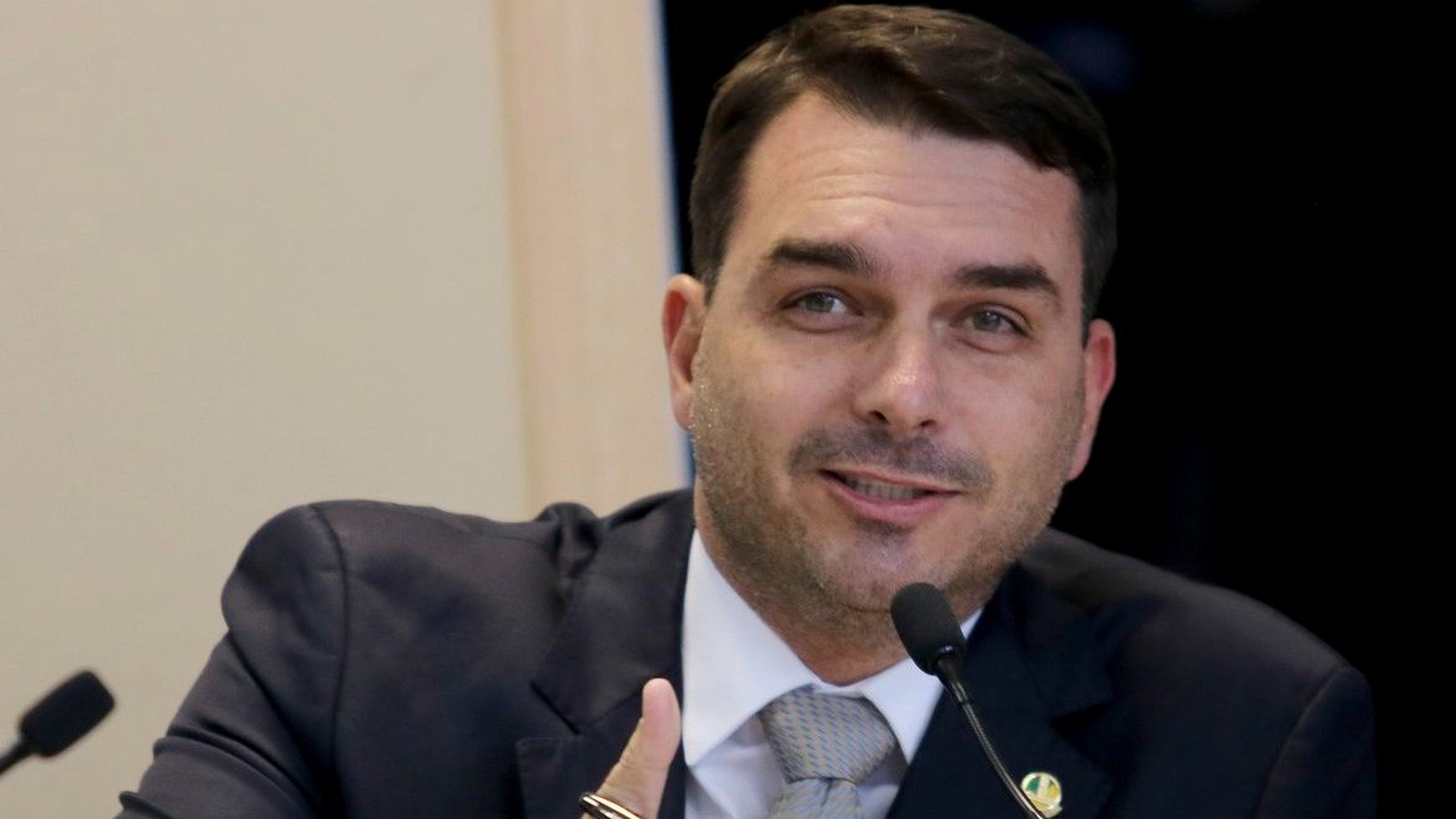 Flávio Bolsonaro de terno e gravata, gesticulando com a mão direita em frente a um microfone