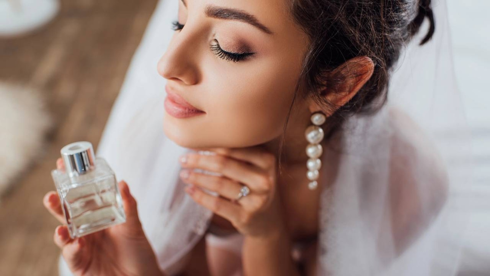 Close-up de mulher com olhos fechados segurando frasco de perfume e usando brincos de pérolas