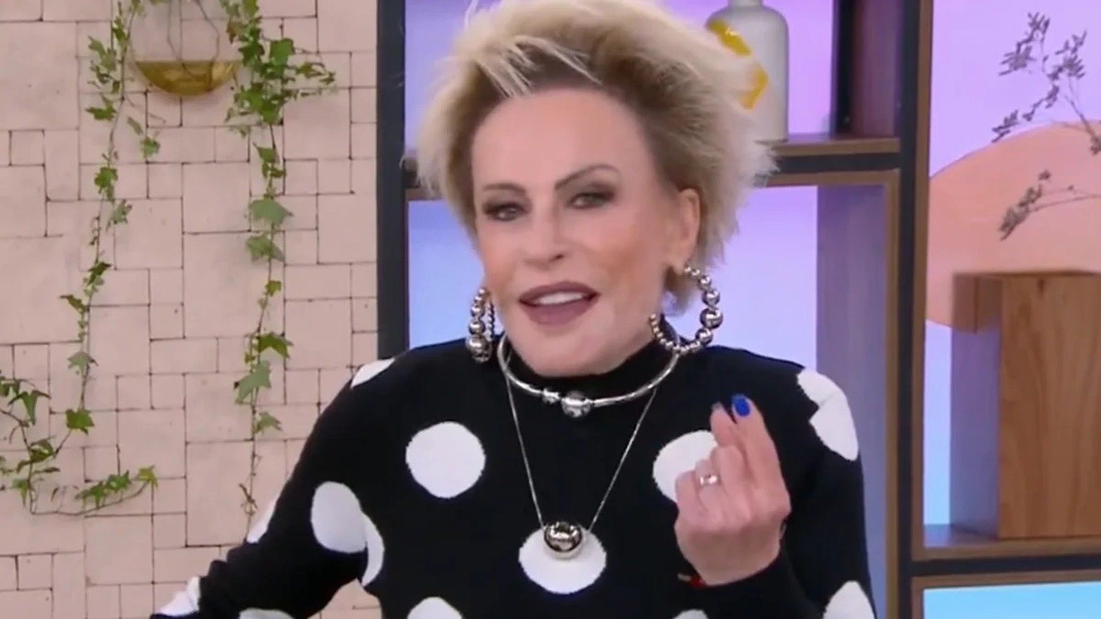 Ana Maria Braga, cabelo loiro curto, blusa preta de bolinhas brancas, gesticula em estúdio