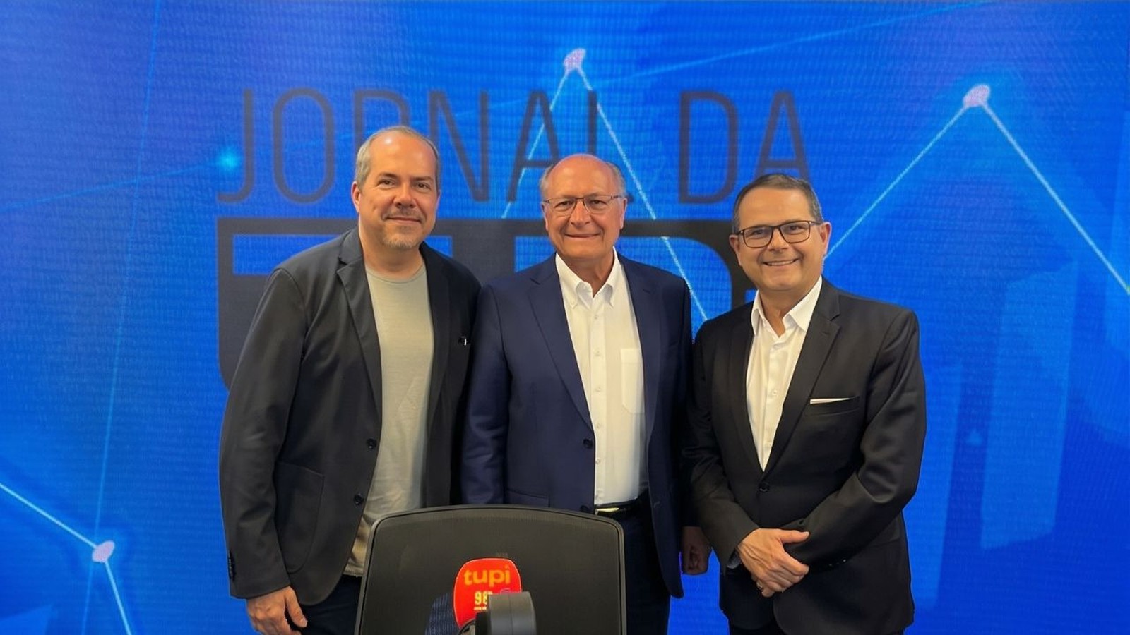 Três homens sorrindo em estúdio de rádio azul com logo "Jornal da Tupi" e "tupi 98"