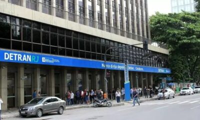Prédio do Detran-RJ com fila de pessoas na calçada, carros estacionados e letreiro 'DETRANRJ' visível