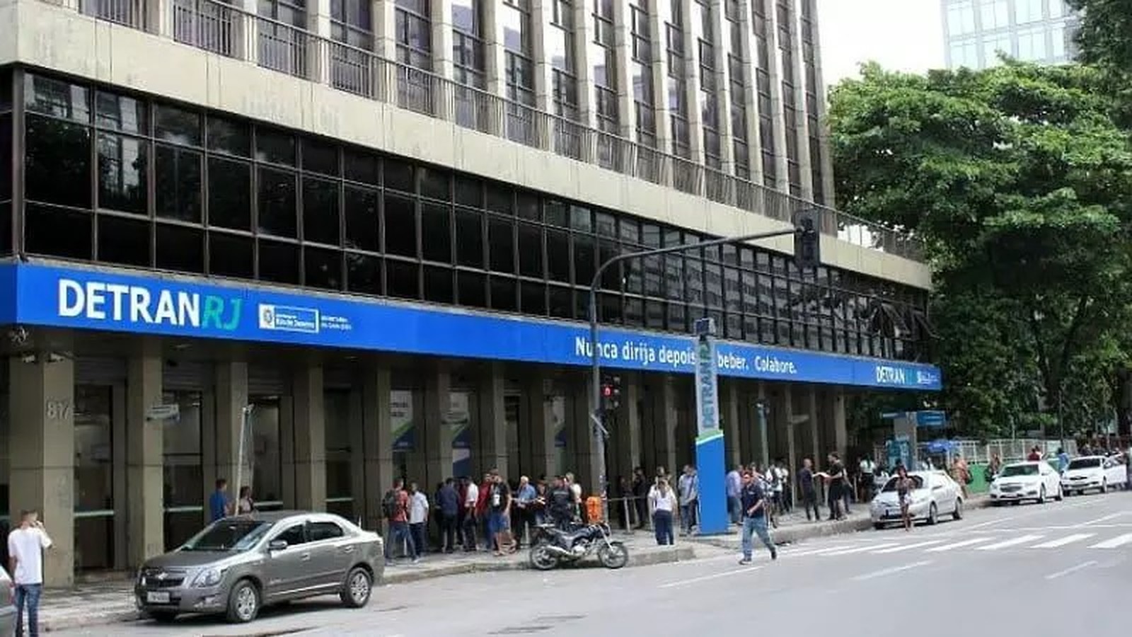 Prédio do Detran-RJ com fila de pessoas na calçada, carros estacionados e letreiro 'DETRANRJ' visível