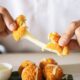 Duas mãos puxam uma bolinha de queijo, mostrando queijo derretido esticando, com mais bolinhas e molho