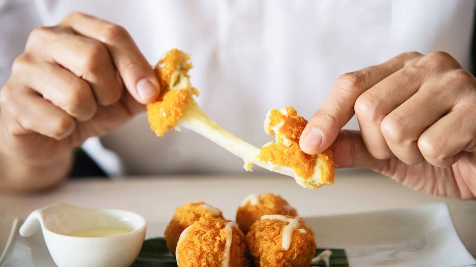 Duas mãos puxam uma bolinha de queijo, mostrando queijo derretido esticando, com mais bolinhas e molho