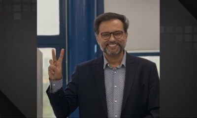 Geraldo Luís sorrindo, usando óculos e barba, fazendo sinal de paz com a mão direita