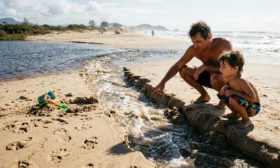 Pai ajuda o filho a cavar um "pequeno rio" entre uma lagoa e o mar durante brincadeira na praia, mas a força da água começa a alargar o canal e transforma a diversão em um possível problema ambiental
