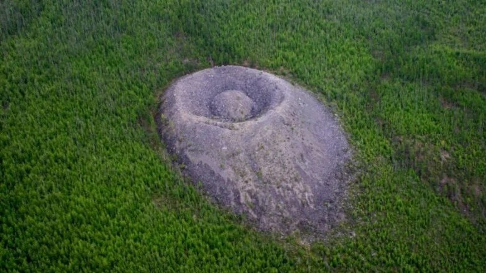 Visão aérea de um monte cônico rochoso cinzento com depressão central, em floresta verde densa