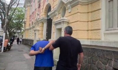 Homem de camisa azul algemado sendo escoltado em calçada por agente com prédio amarelo atrás