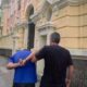 Homem de camisa azul algemado sendo escoltado em calçada por agente com prédio amarelo atrás