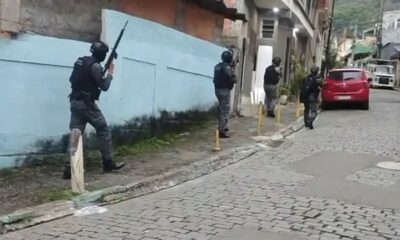 Quatro policiais táticos com fuzis em rua de paralelepípedos, com casas e carros