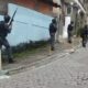 Quatro policiais táticos com fuzis em rua de paralelepípedos, com casas e carros