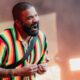 Homem negro barbudo com camisa listrada colorida cantando em microfone headset no palco