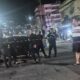 Policiais isolam carro preto à noite em rua, com fita amarela e mancha escura no chão