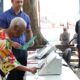 Mulher idosa vota em urna eletrônica, homem com pasta, banner 'VOTO NA DEMOCRACIA'