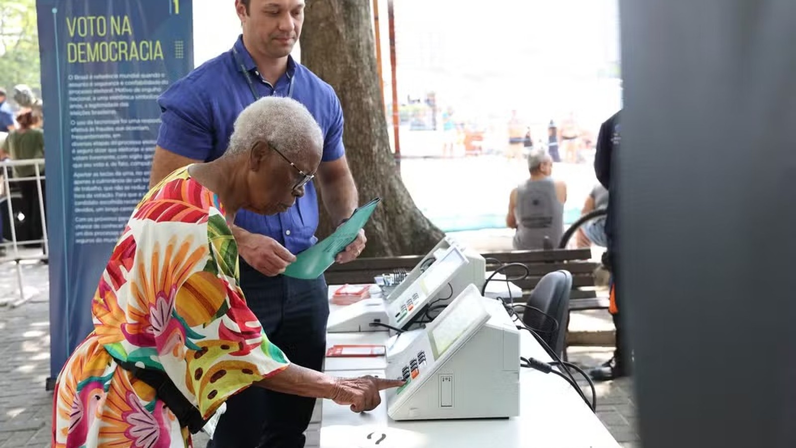 Mulher idosa vota em urna eletrônica, homem com pasta, banner 'VOTO NA DEMOCRACIA'