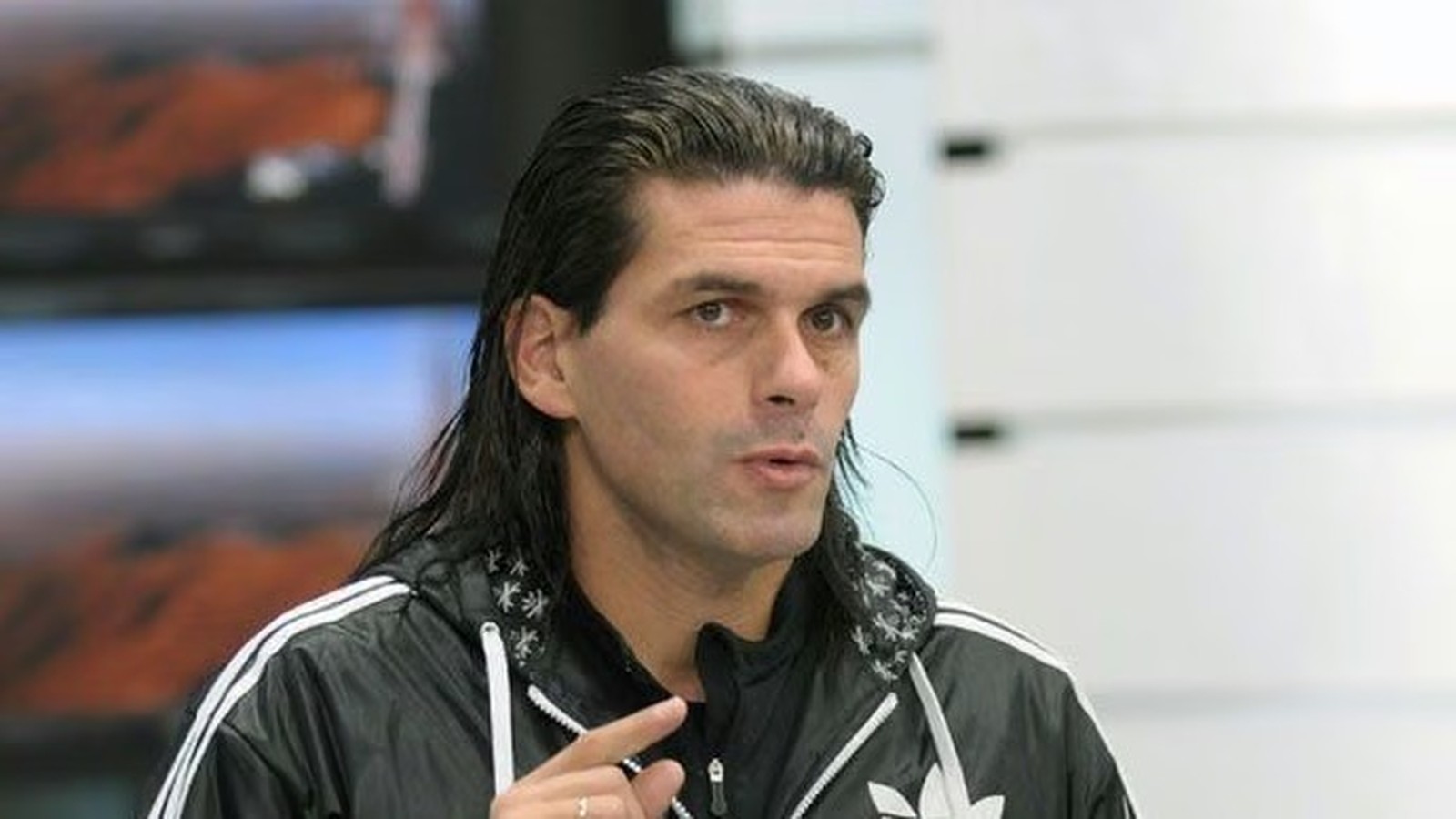 Homem de cabelo comprido e jaqueta preta Adidas gesticula enquanto fala
