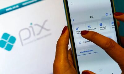 Mãos com unhas vermelhas usando smartphone com aplicativo Pix, e logo Pix no fundo