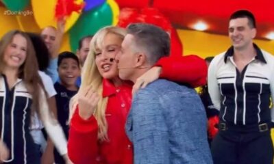 Homem beija mulher loira em palco, plateia sorridente, fundo colorido de programa de TV