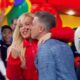 Homem beija mulher loira em palco, plateia sorridente, fundo colorido de programa de TV