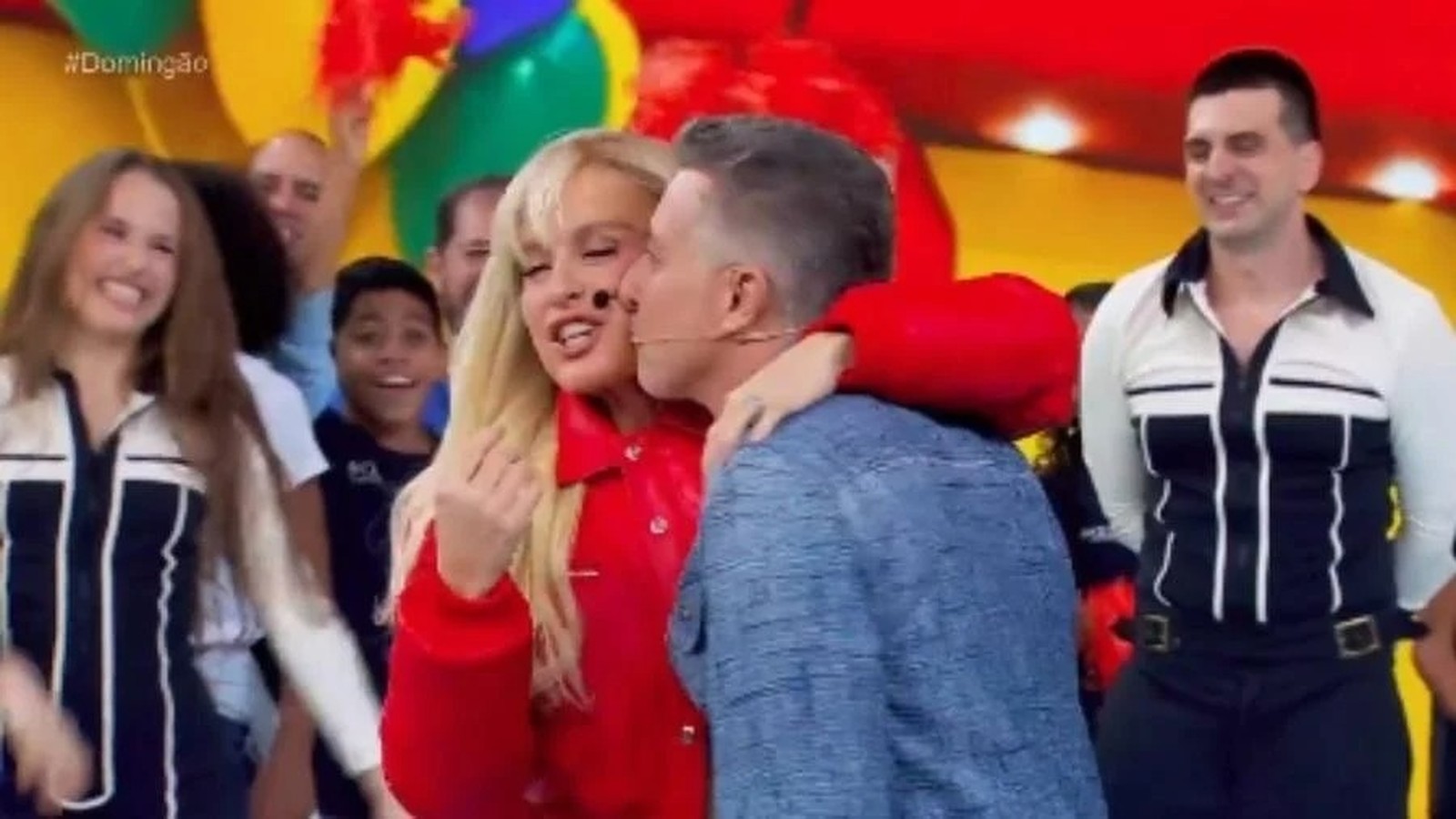 Homem beija mulher loira em palco, plateia sorridente, fundo colorido de programa de TV
