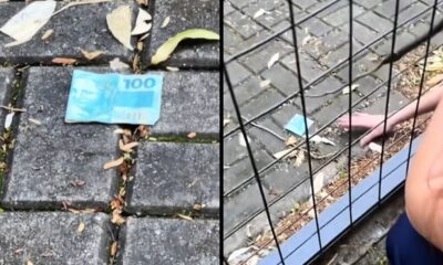 Uma nota de R$ 100 em chão de pedra e uma mão a alcançando por grade preta.