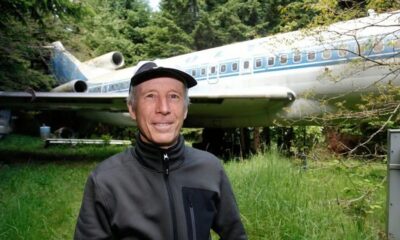 Homem sorridente com boné e jaqueta em frente a avião Boeing 727 na floresta