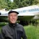Homem sorridente com boné e jaqueta em frente a avião Boeing 727 na floresta