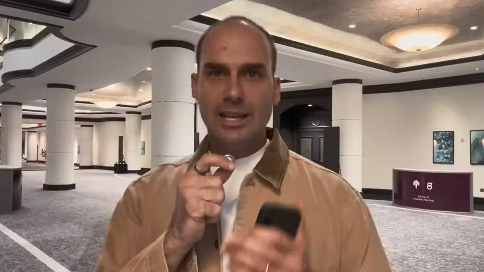 Homem com jaqueta marrom e anel segura celular, gesticulando em saguão com pilares brancos