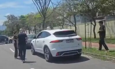 SUV branco e carro escuro em rua com três pessoas e árvores em dia ensolarado
