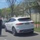 SUV branco e carro escuro em rua com três pessoas e árvores em dia ensolarado