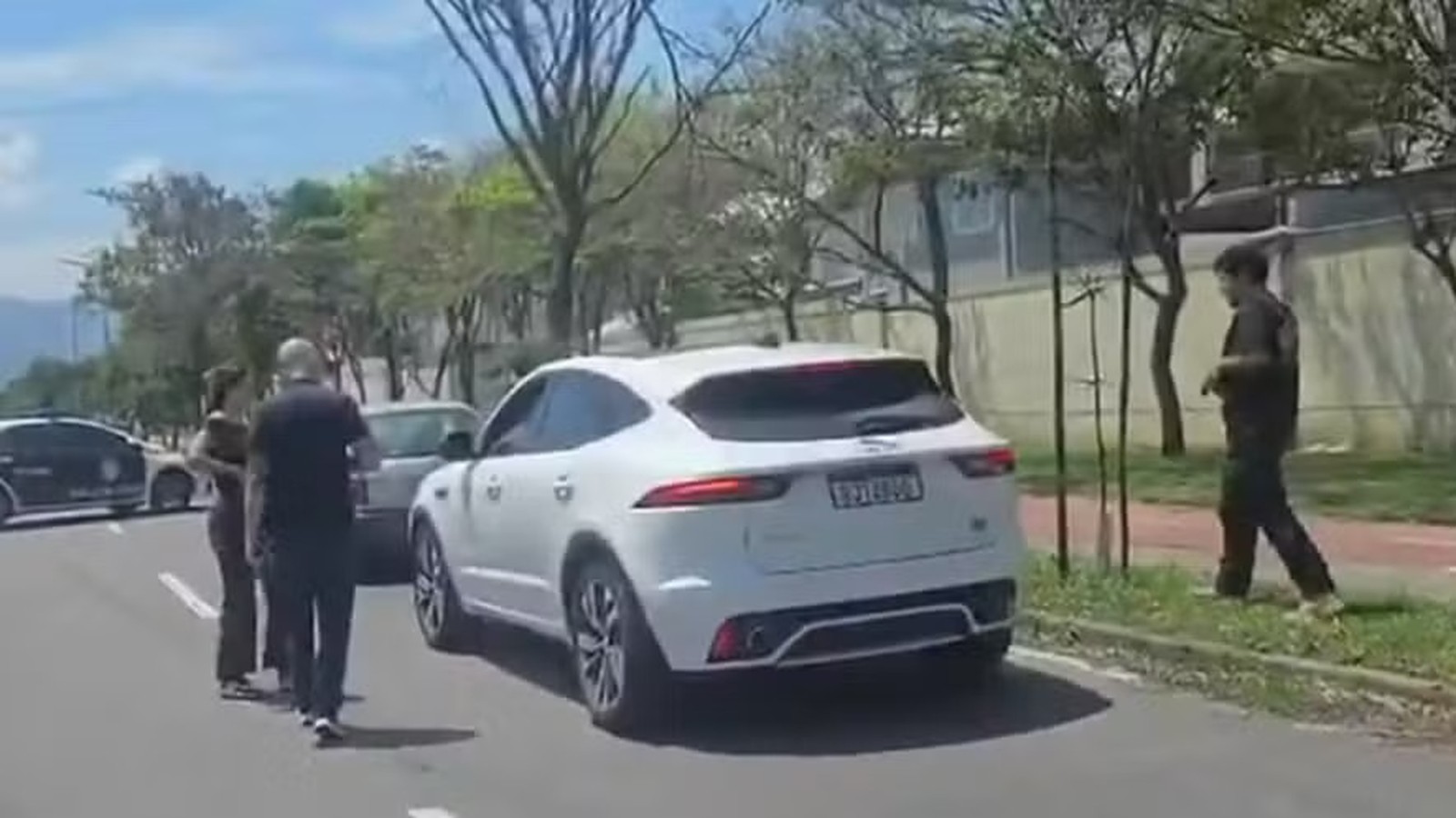 SUV branco e carro escuro em rua com três pessoas e árvores em dia ensolarado